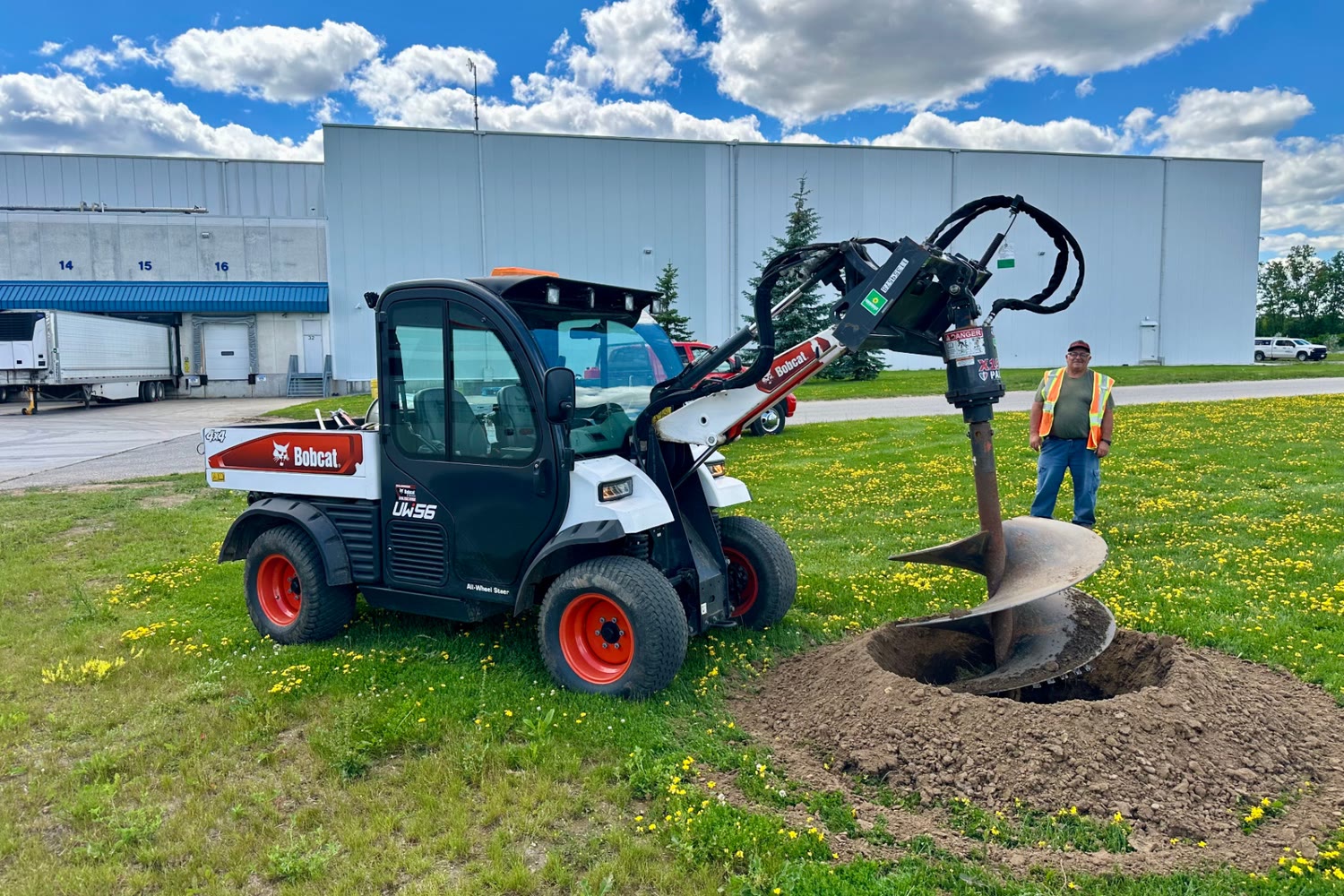 Bobcat UW56 with a tree-planting auger at an industrial Brantford property, with a crew member in a high-visibility vest nearby.