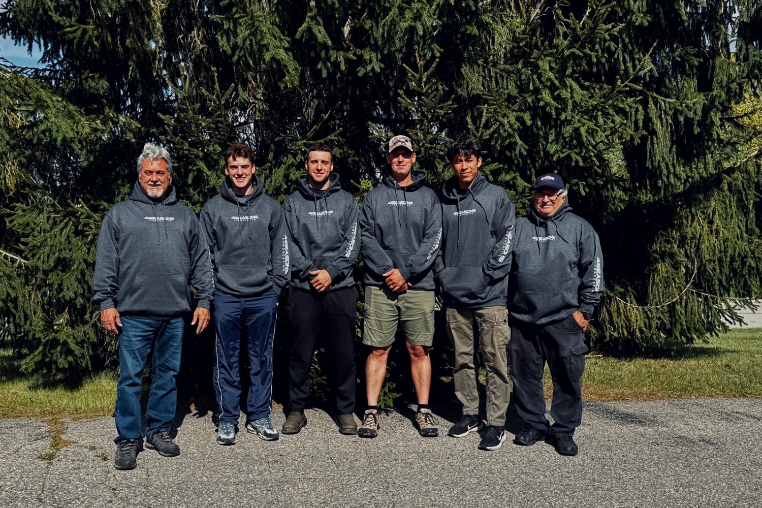Six uniformed 4 Seasons Landscaping crew members standing together in front of a row of evergreens.