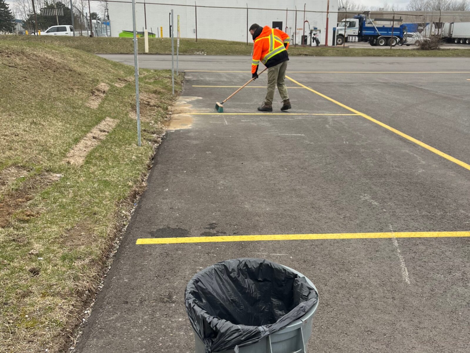 Crew member in a high-visibility jacket sweeping a commercial parking lot beside lined stalls and waste bins in Brantford.
