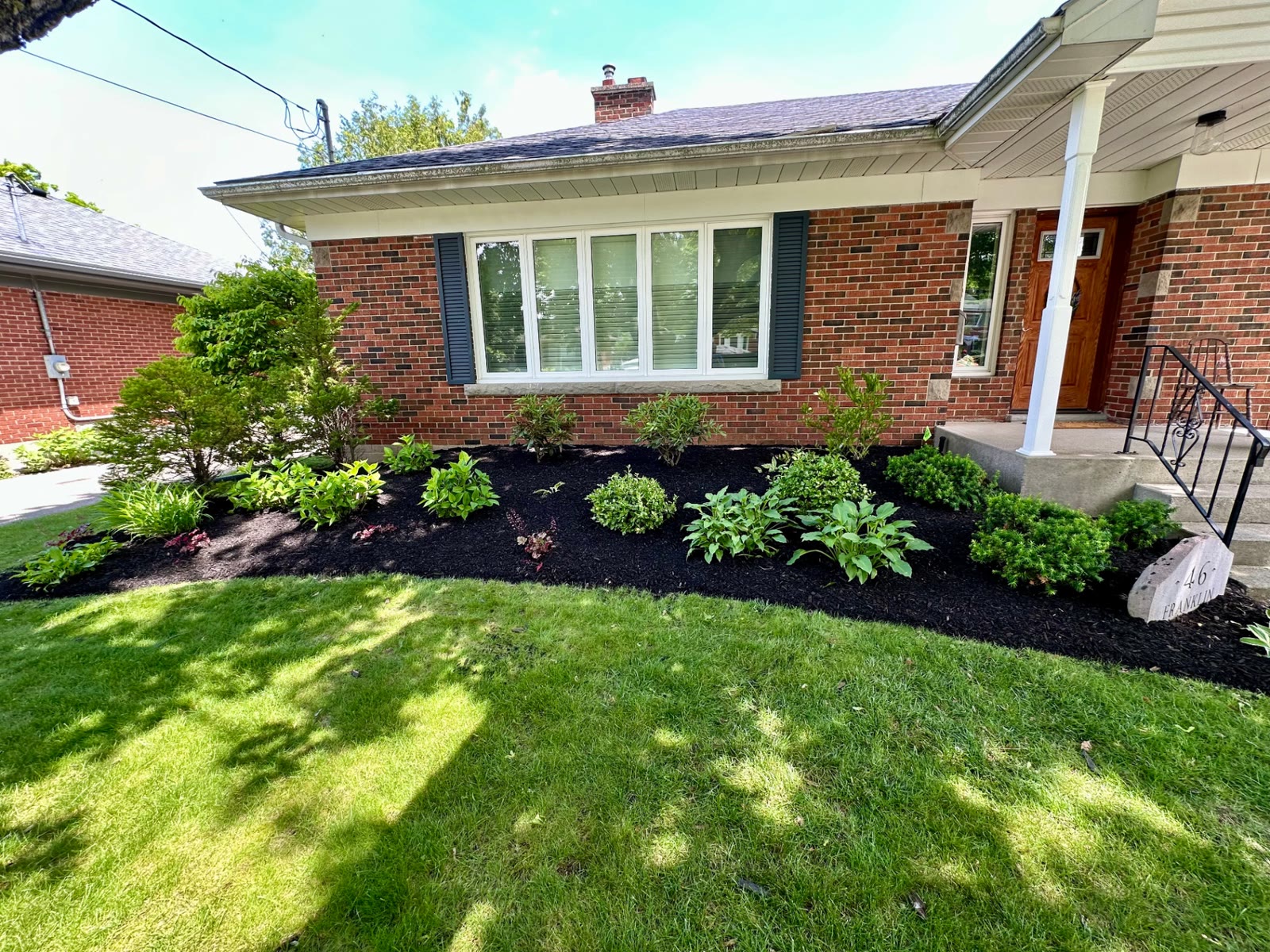 Curved front bed with hostas, boxwood and flowering shrubs in dark mulch next to a house number stone.